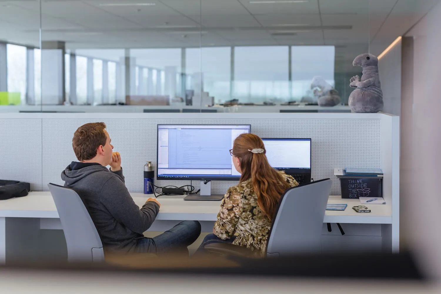 Two colleagues are seated side by side in a modern office, collaborating while looking at a large computer monitor and a laptop